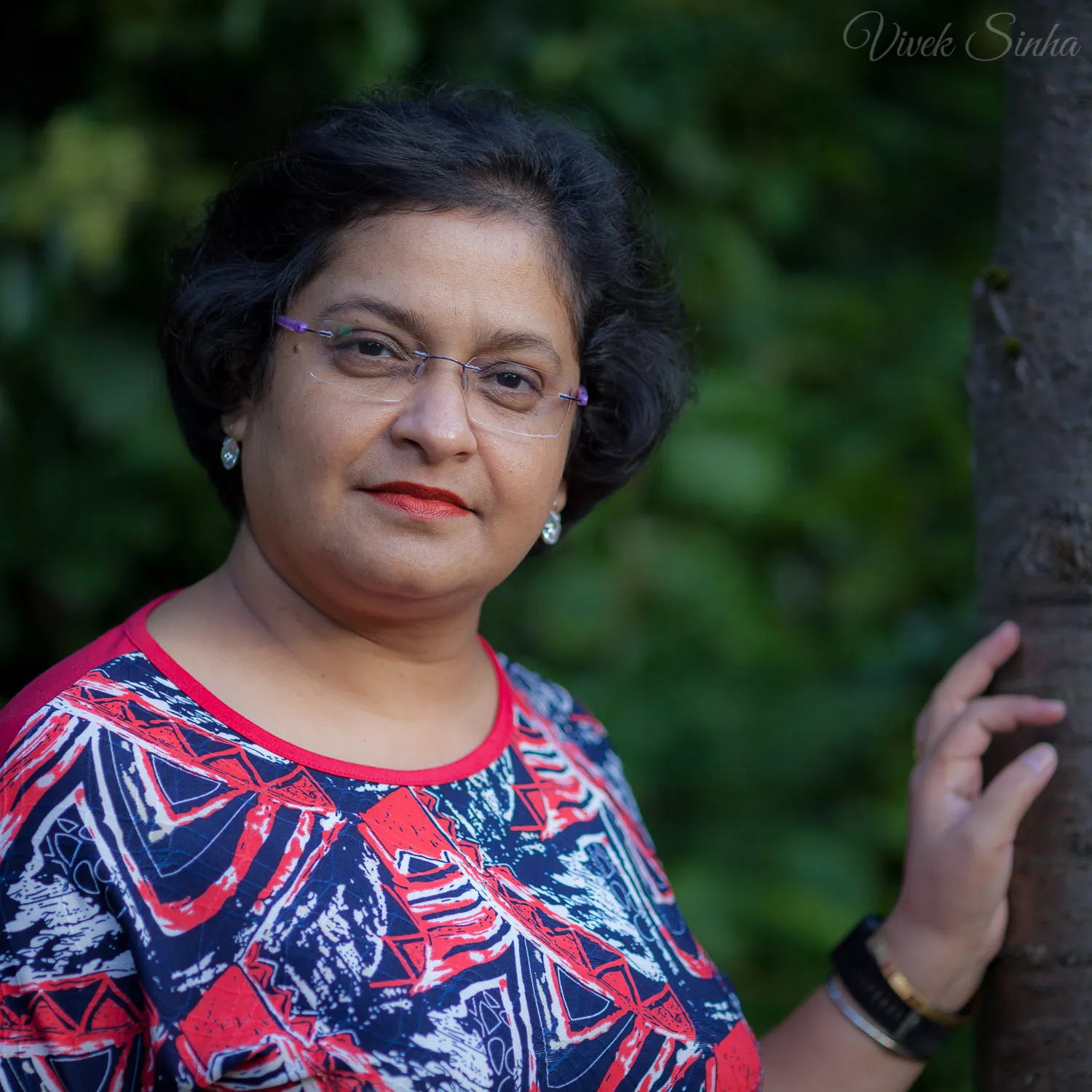 A woman with short dark hair and rimless glasses smiles slightly, wearing a red, white, and navy patterned top, resting one hand on a tree trunk with dark green foliage behind her.