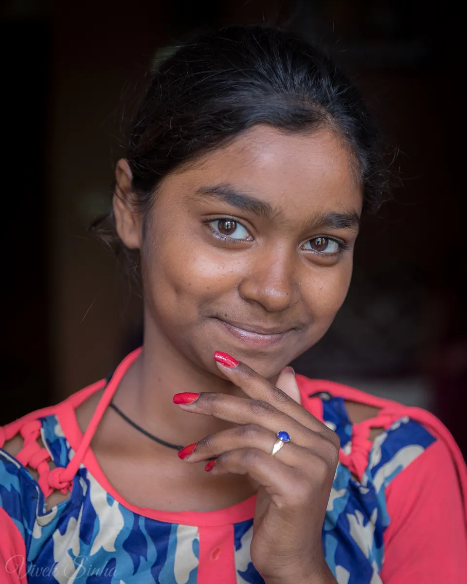 A young girl with dark hair and a gentle smile rests red-painted fingernails against her chin, wearing a pink-and-blue patterned top against a dark interior background.