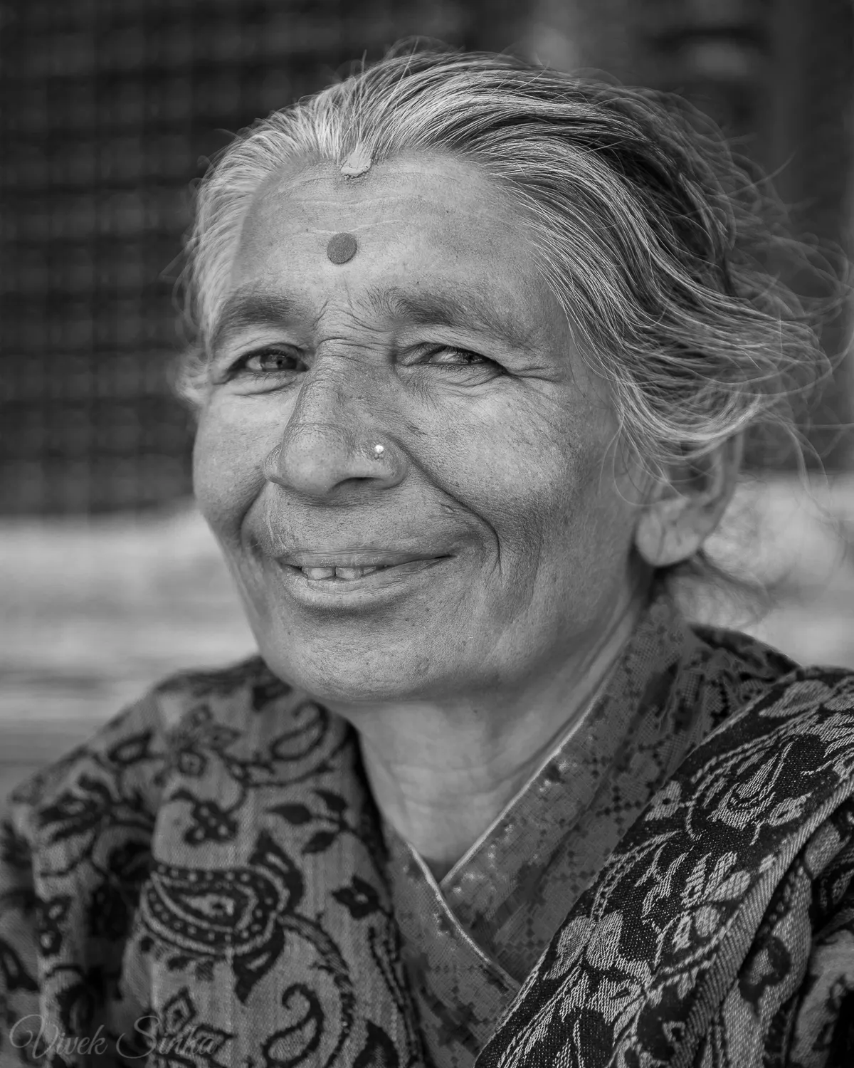 Black-and-white portrait of a smiling older woman in Kathmandu with grey-streaked hair, a bindi, and a nose stud, wearing a patterned shawl.