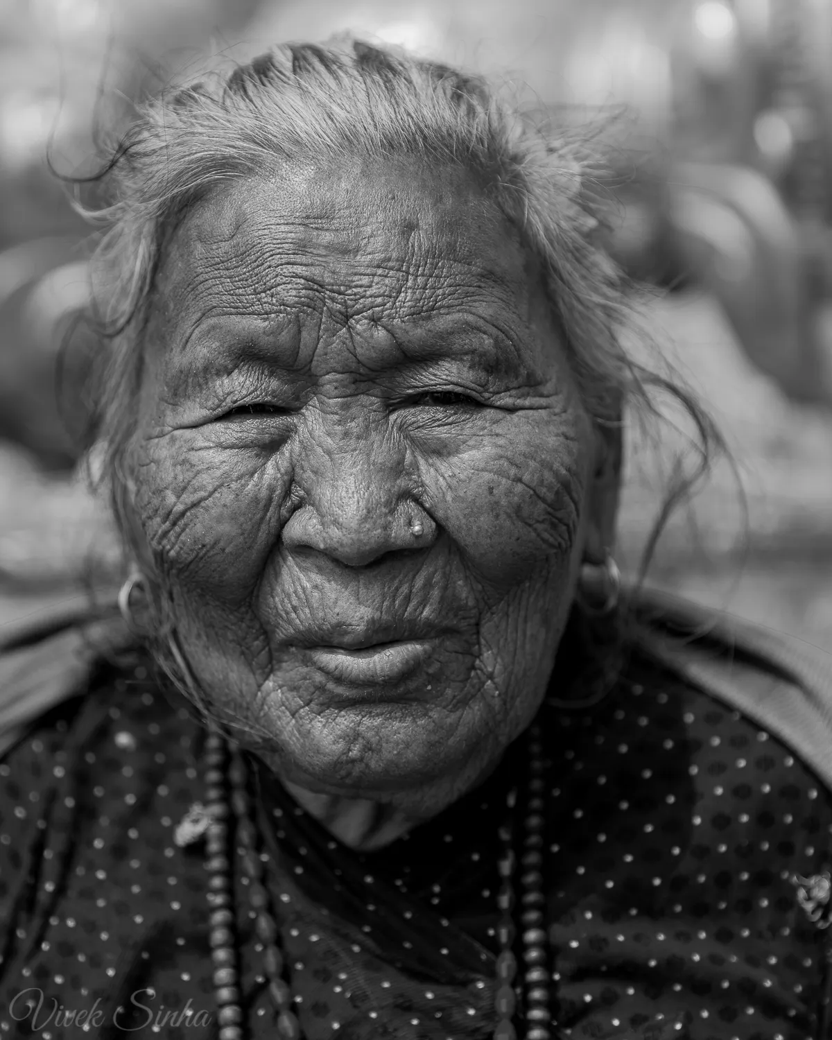 Black-and-white close-up of an elderly woman in Kathmandu with deeply wrinkled skin, wind-blown grey hair, a nose stud, and beaded necklaces, looking into the camera.