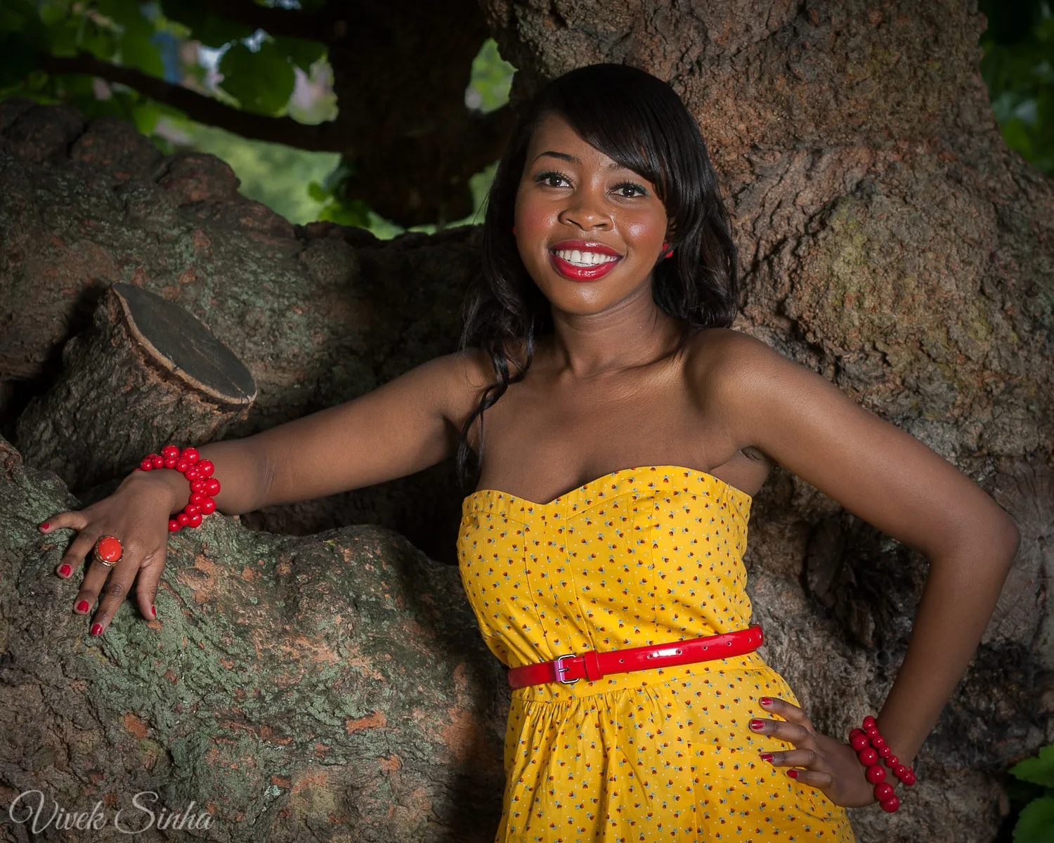 A smiling young woman in a strapless yellow polka-dot dress with a red belt and red bead bracelets poses with one arm on the gnarled trunk of a large tree.