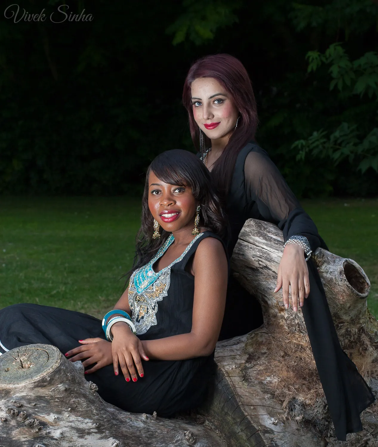 Two women in black outfits with jewelled details pose on a large fallen tree trunk in a park, one seated in front and the other leaning behind her against dark green foliage.