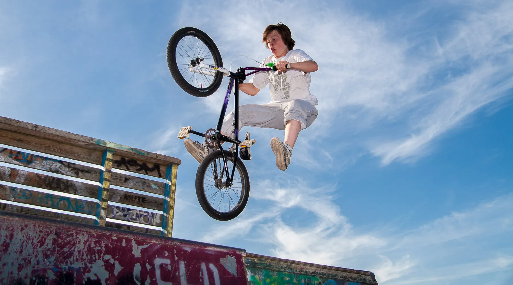 A boy in a white t-shirt flies through the air above a graffiti-covered skate ramp, his BMX bike tilted with the front wheel high while his feet leave the pedals against a blue sky.