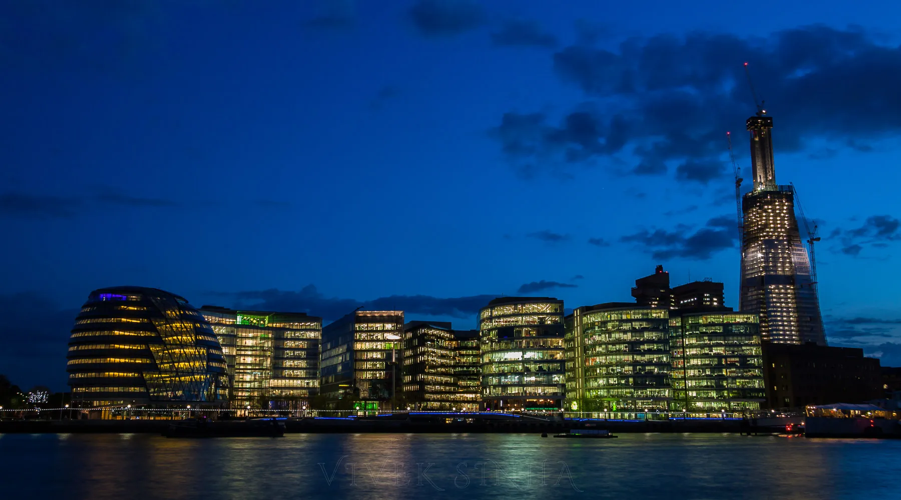 A deep-blue dusk skyline along the Thames with the unfinished Shard skyscraper and construction cranes towering over illuminated riverside office buildings, including the rounded City Hall.