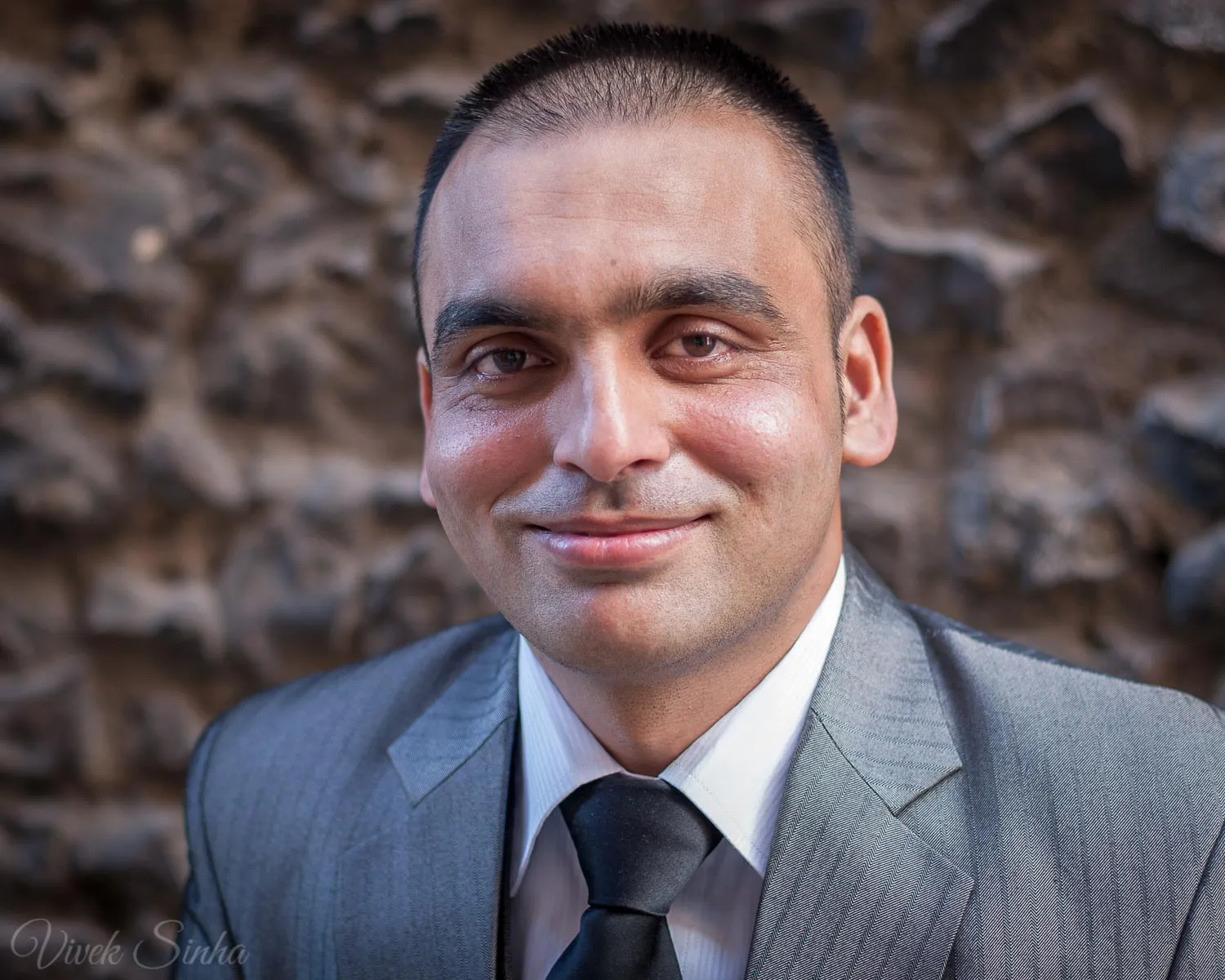 A man with closely cropped hair smiles slightly, wearing a grey suit, white shirt, and black tie in front of a blurred dark stone wall.
