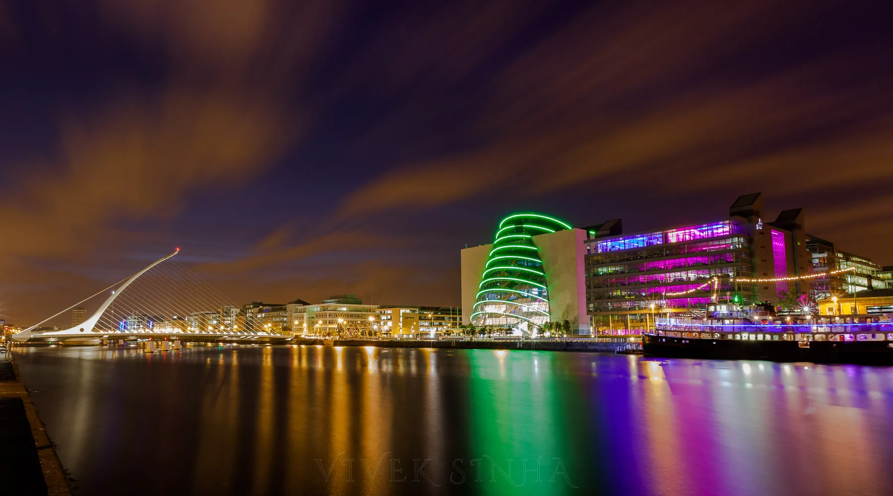 A night view of Dublin's River Liffey with the cable-stayed Samuel Beckett Bridge on the left and buildings lit in green, pink, and purple reflecting in the water.