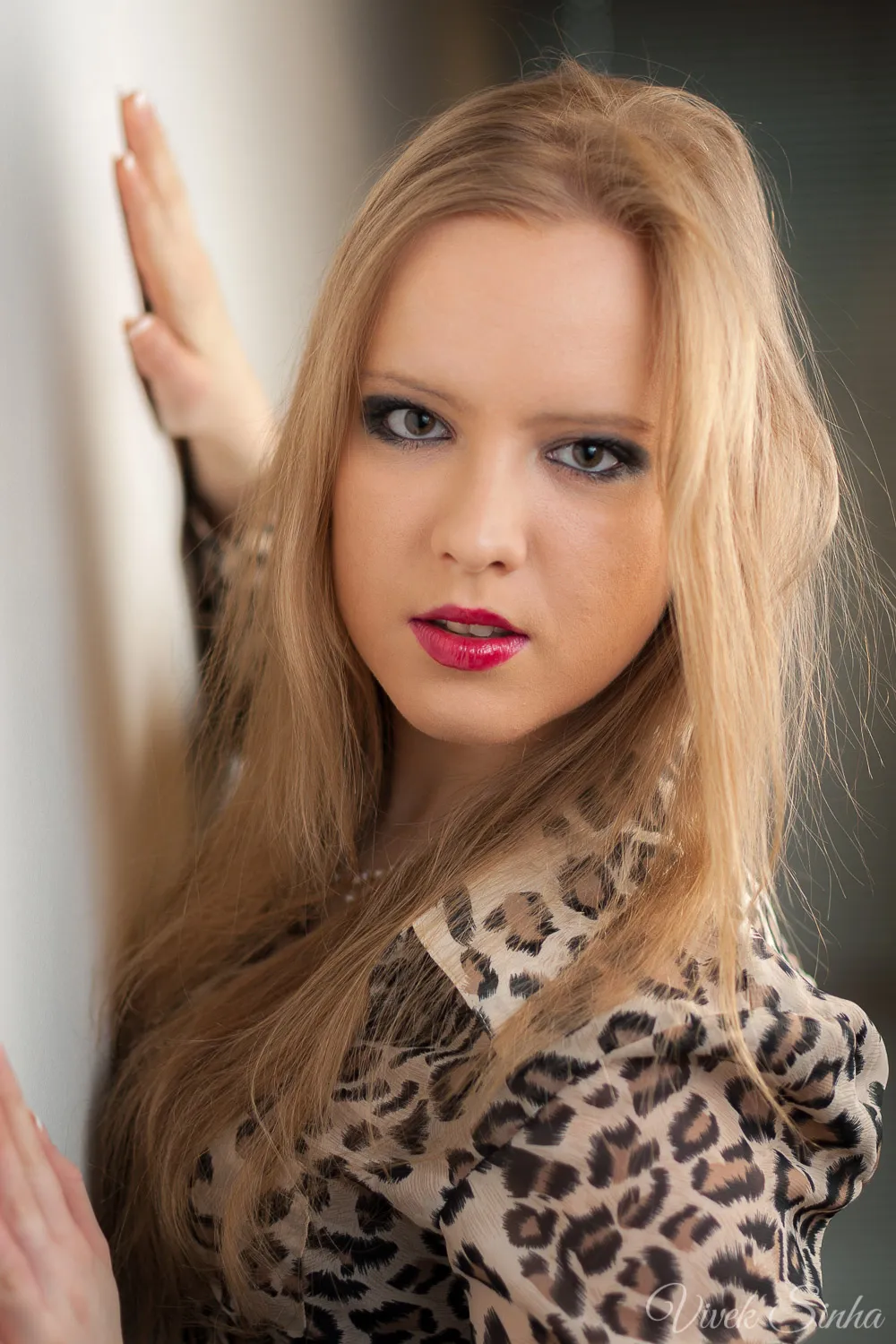 A young woman with long blonde hair, dark eye makeup, and red lipstick wears a leopard-print blouse and rests one raised hand against a pale wall indoors.